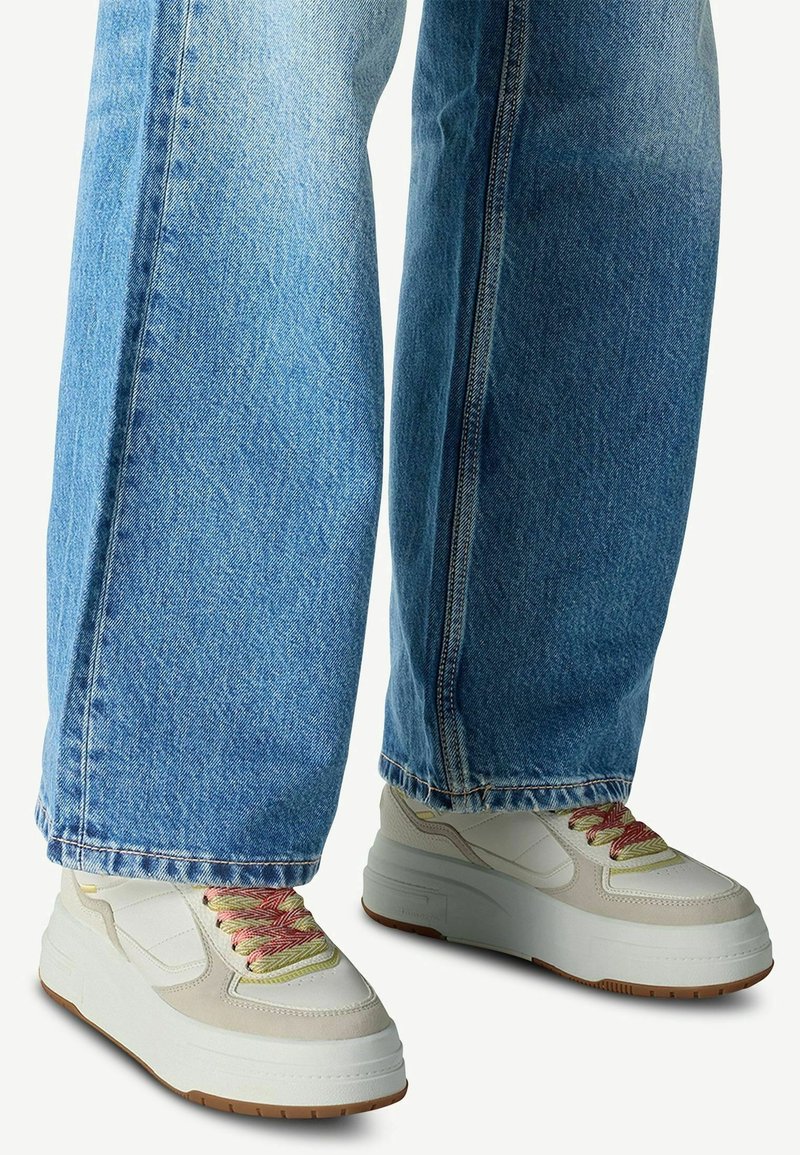 Jambes portant un jean bleu clair à jambes larges et des baskets blanches à plateforme avec des lacets roses et jaunes sur un fond uni.