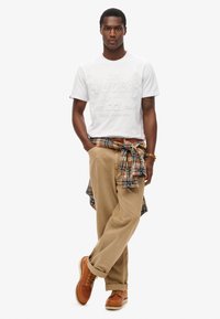 Hvid kortærmet T-shirt med præget logo, parret med lysebrune bukser. En ternet skjorte bundet om livet og brune snørebåndsstøvler.