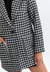 Svartrutigt houndstooth-mönstrad kavaj med slagkrage, enkel knappstängning och sidofickor. Strukturerat tyg.