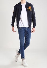 Chaqueta bomber de la marina con parches, camisa blanca, vaqueros azules y zapatillas blancas. La chaqueta presenta un cierre de cremallera y puños acanalados.
