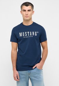 Mārī zila īsām piedurknēm T-kreklā, kas izgatavots no kokvilnas, ar baltu logo "MUSTANG" un tekstu. Modelim mugurā ir gaiši zili džinsi.