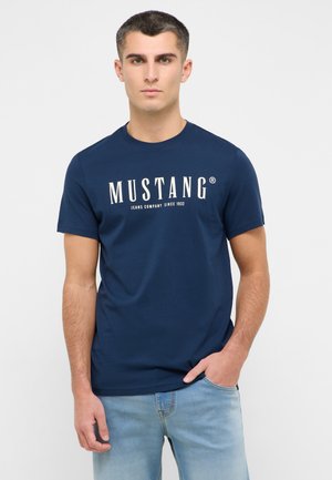 Granatowa koszulka z krótkim rękawem, wykonana z bawełny, z białym logo "MUSTANG" oraz napisem. Model nosi jasnoniebieskie dżinsy.
