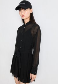 Črna šifonska obleka s ovratnikom, gumbi na sprednji strani, dolgimi rokavi in zavezanim pasom. Ima plisirano krilo in srebrne gumbnice.