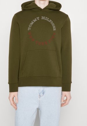 Oliwkowo-zielona bluza z kapturem z przednią kieszenią i okrągłym napisem „Tommy Hilfiger” oraz „New York City”, noszona z jasnoniebieskimi dżinsami.