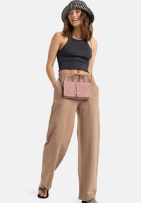 Světle růžová crossbody kabelka s klopou, obdélníkový tvar, hladký povrch. Modelka má na sobě tmavý zkrácený top, béžové široké kalhoty a kostkovanou čepici.