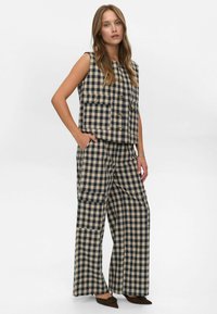 Zweiteiliges Outfit im beigefarbenen und marineblauen Vichykaro-Muster; ärmelloses Oberteil mit zwei Taschen; weite Hose mit Seitentaschen; flache Schuhe.