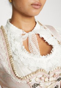 Femme portant un chemisier en dentelle blanche à col festonné et nœud, associé à un corset floral rose pâle avec des détails de ruban et de fermoirs métalliques.