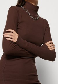 Brun ribbad polotröja med långa ärmar och slim fit, prydd med ett silverkedjehalsband som accentdetalj.