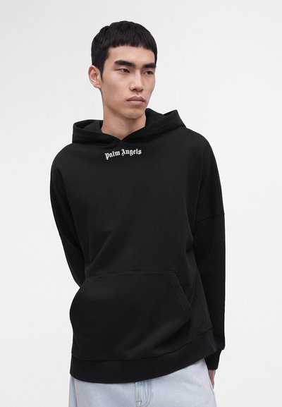 Jeune homme portant un sweat à capuche noir Palm Angels avec une poche avant et un jean bleu clair, debout devant un fond blanc uni.