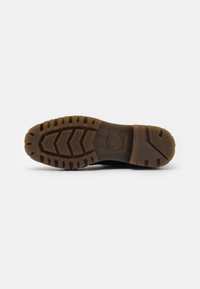 Semelle de chaussure noire avec un motif texturé et des rainures. Le matériau est en caoutchouc avec une semelle intermédiaire marron. Présente un logo circulaire au niveau du talon.