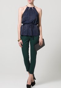 Marinblå halterneck-top med rynkad halsringning, knytmidja; gröna åtsittande byxor; svart kuvertväska; högklackade skor. Slät textur, avslappnad design.