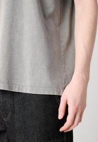 Gray cotton t-shirt with a relaxed fit, featuring subtle texture and embroidered "MN" detail at the hem, paired with dark denim jeans.
