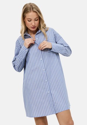 Blond vrouw die een blauw-wit verticaal gestreepte lange mouwen overhemdjurk losknoopt tegen een effen lichte achtergrond.