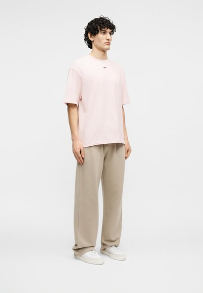 Νέος άνδρας με σγουρά μαλλιά φοράει ένα μεγάλο μπλουζάκι σε απαλό ροζ χρώμα, μπεζ παντελόνι με φαρδιά μπατζάκια και λευκά αθλητικά παπούτσια, στέκεται μπροστά σε απλό λευκό φόντο.