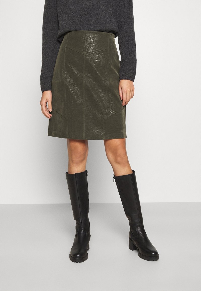 Jupe A-line en simili cuir de couleur vert olive avec des coutures avant et une texture lisse, associée à des bottes en cuir noires montantes.