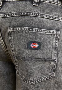 Pantalones de mezclilla negra con un bolsillo trasero que presenta una etiqueta rectangular en rojo, amarillo y azul con el logo "Dickies"; tejido texturizado visible.
