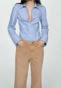 Camicia a righe azzurro chiaro con bottoni sul davanti e colletto, abbinata a jeans beige a vita alta, che mettono in risalto una silhouette aderente.