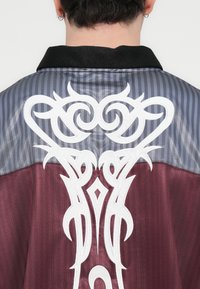 Πουκάμισο με γιακά σε γκρι και μαροον ύφασμα με ριγέ σχέδιο. Διαθέτει λευκή φυλετική κεντητική στο πίσω μέρος, προσθέτοντας υφή και λεπτομέρειες.