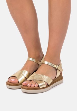 Piedi che indossano sandali in pelle metallizzata dorata con cinturini incrociati, fibbia alla caviglia e suola beige imbottita su uno sfondo grigio chiaro.