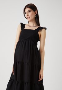 Rochie de maternitate neagră cu volane pe bretele, corp smockat și o siluetă fluidă. Fabricată dintr-un material ușor pentru confort.