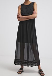Svart ärmlös maxiklänning med blommig spetsdetalj vid bysten och nederkanten, med en genomskinlig överdel och scalloped-kant.