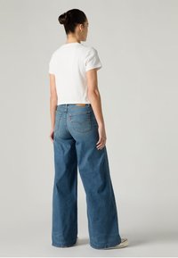 Τζιν με φαρδιά πόδια σε μεσαίο μπλε ντένιμ με ψηλή μέση. Συνδυασμένο με ένα κοντό λευκό T-shirt. Ελάχιστα μεταλλικά στοιχεία και κανένα ορατό μοτίβο.