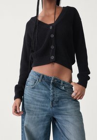 Zwart gebreid kort vest met V-hals en vijf knoopsluitingen, gecombineerd met lichtblauwe losse jeans. Gestructureerde stof.