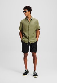 Camicia verde oliva a maniche corte con bottoni, realizzata in tessuto leggero, abbinata a pantaloni corti neri e sneakers nere con dettagli bianchi.