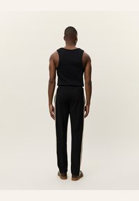 Schwarzes, ärmelloses Tanktop, kombiniert mit schwarzen Hosen, die helle beige Seitenstreifen haben, und von hinten eine schlanke Passform und weiche Textur zeigen.