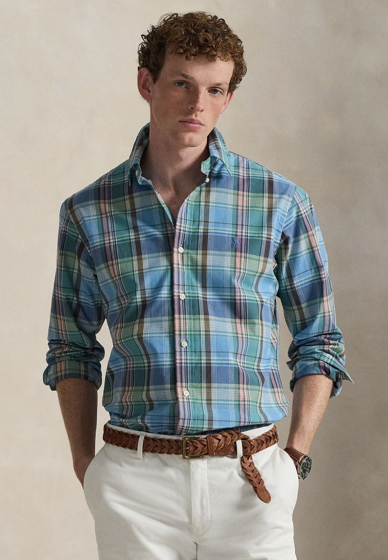 Men's plaid shirt in blue, green, pink, and purple tones; button-down collar, long sleeves rolled up, worn with white trousers and a brown braided belt.
