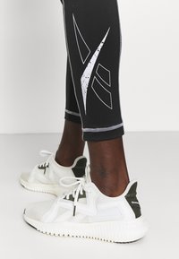 Czarne legginsy z białymi graficznymi akcentami zestawione z białymi trampkami z teksturowaną siateczką i zielonymi akcentami.