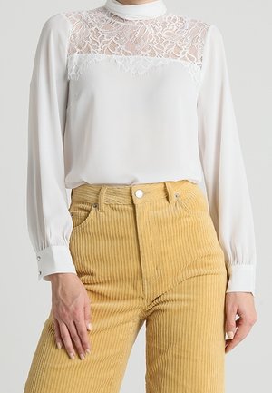 Witte blouse met lange mouwen en kanten inzetstuk op het bovenstuk, gecombineerd met mosterdgele corduroy broek met hoge taille, gedragen door een staande persoon.
