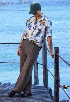 Junger Mann mit einem Blumenshirt, brauner Hose, grünem Cap und Sonnenbrille lehnt an einem hölzernen Geländer am blauen Wasser.