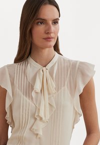 Light beige blouse with ruffled sleeves, vertical pinstripes, round collar, and a tied bow at the neck. Sheer, flowing fabric adds texture.