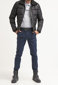 Doudoune noire avec un col bordé de fourrure, polo gris, chinos bleus et bottines noires avec des accents de boucle.