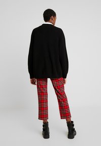 Černý pletený svetr s oversized střihem, kombinovaný s červenými kostkovanými kalhotami s černými a žlutými pruhy, nošený s černými kotníkovými botami.