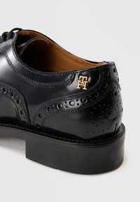Black leather dress shoe featuring brogue detailing, a smooth finish, and a gold logo accent. The shoe has a sturdy black rubber sole.