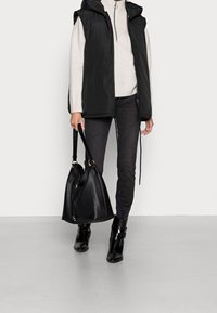 Schwarze gepolsterte Weste mit Kapuze, grauer Pullover, dunkle Jeans und schwarze Stiefeletten; hält eine große schwarze Handtasche mit goldenen Hardware-Akzenten.