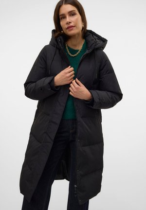Femme portant un long manteau noir matelassé avec capuche, un pull vert, un jean foncé et un collier en or, debout devant un fond blanc uni.