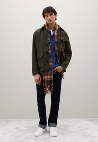 Young man wearing an olive green jacket, blue shirt, dark jeans with cuffs, white sneakers, and a red plaid scarf standing against a plain background.