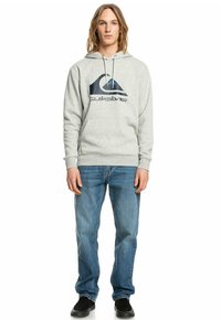 Grå hoodie med en stor marinblå Quiksilver-logotyp, med en kenguruficka och justerbara dragskor. Matchas med blå jeans och svarta skor.