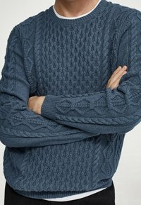 Pull bleu en maille torsadée avec un col rond. Présente des motifs texturés sur les manches et le corps, des poignets et un ourlet côtelés.