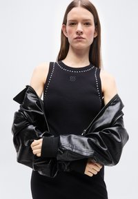 Γυναίκα με ίσια καστανά μαλλιά που φοράει μαύρο αμάνικο τοπ με λεπτομέρειες με τρουκς και μαύρο δερμάτινο μπουφάν που φοριέται μερικώς ανοιχτό.