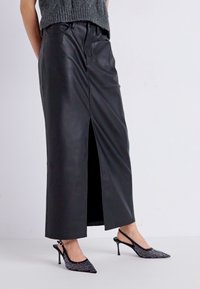 Jupe maxi en cuir noir avec une fente à l'avant, présentant une texture lisse et une taille haute. Associée à des escarpins slingback noirs à talons aiguilles ornés de clous.