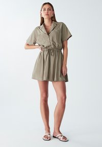 Giovane donna che indossa un vestito khaki a maniche corte con bottoni e una cintura con cordoncino, abbinato a sandali bianchi con cinturini piatti, in piedi contro uno sfondo neutro.