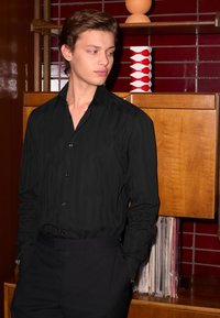 Schwarzes gestreiftes Hemd mit Kragen, geknöpften Manschetten und figurbetontem Schnitt, kombiniert mit maßgeschneiderten schwarzen Hosen vor einem hölzernen Hintergrund.