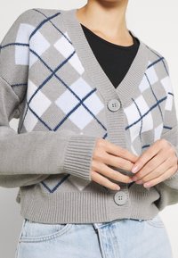 Harmaa neuletakki, jossa on timanttikuvio valkoisena ja sinisenä, V-pääntie, etupainikehä ja ribbireunat. Pehmeä neuletekstuuri.