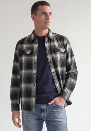 Mand står med den ene hånd i lommen iført sort og gråternet skjorte over navy t-shirt og lyseblå jeans mod en ensfarvet baggrund.