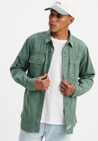 Camisa verde de botones con dos bolsillos en el pecho, mangas largas y un corte holgado. Fabricada en una tela suave con un acabado mate. Camiseta blanca debajo.
