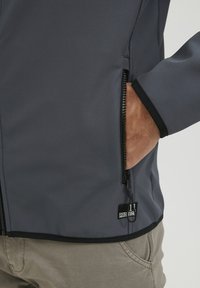 Gray softshell jacket featuring a zippered pocket, contrasting black accents, and a textured fabric. Includes a logo tag on the hem.